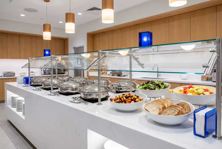 A white stone buffet counter with steel chafing dishes under glass guards, bowls of salad, roasted vegetables, bread and fruit, oak panels behind