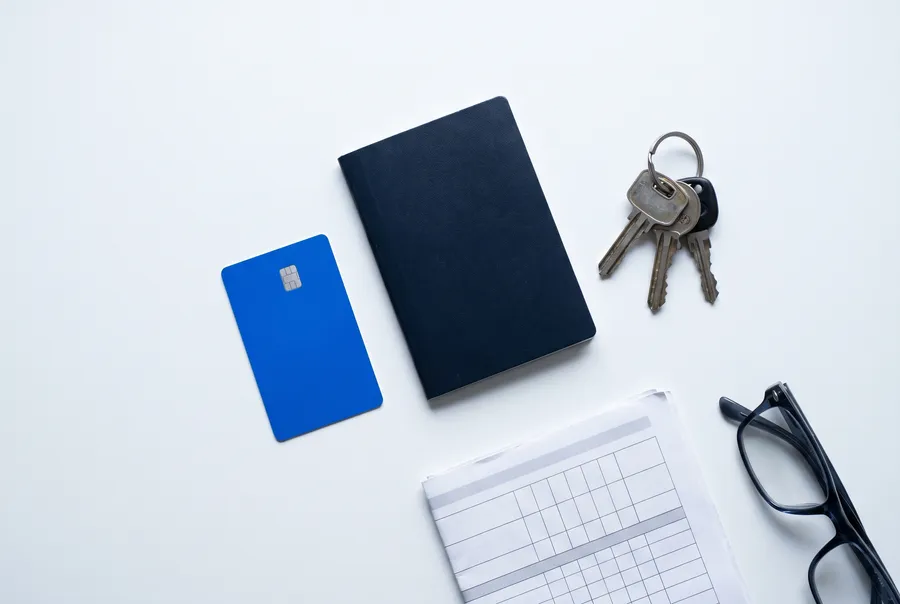 A flat lay on a white desk: a blank electric-blue plastic card with a chip, a closed navy passport-style booklet, a ring of keys, a folded timetable and reading glasses